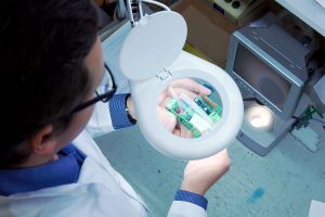 A man in a white robe and glasses checks the electrical Board for defects through a magnifying glass. Quality control department in the production. manufacturing in a high tech factory.